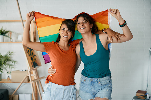 Young happy lesbian couple having fun and holding rainbow flag at home while looking at camera.