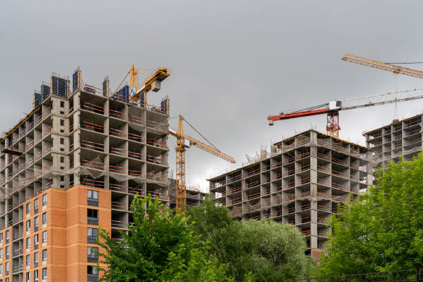 Monolithic concrete frame of apartment building under construction with partially attached ready-made facade cladding blocks. Construction cranes and unfinished residential buildings against grey sky. stock photo