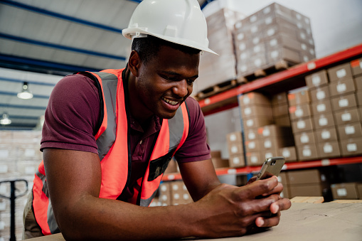 African American male worker texting on cellular device chatting to overseas family while working in warehouse. High quality photo