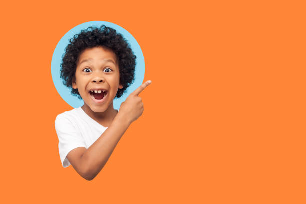 Wow look, advertise here! Portrait of amazed cute little boy with curly hair pointing to empty place Wow look advertise here! Portrait of amazed little boy with curly hair pointing to empty place on background showing copy space for promotional ad. indoor isolated in a round hole on orange background primary age child stock pictures, royalty-free photos & images