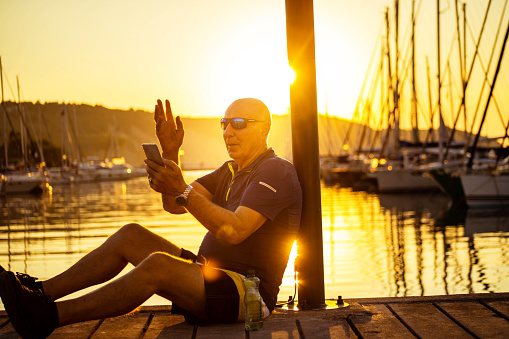 Modern senior man, sitting on the wooden pier in the marina, and having a video call, while enjoying the majestic sunset