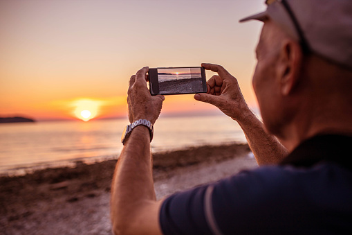 Joyful senior male tourist, taking a photo of sunset above the sea, on the beach with his mobile phone, during his sea vacation
