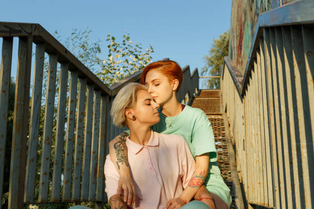 Adorable young lesbian couple in stylish clothes sits together on metal stairs under clear blue sky at sunset in summer evening