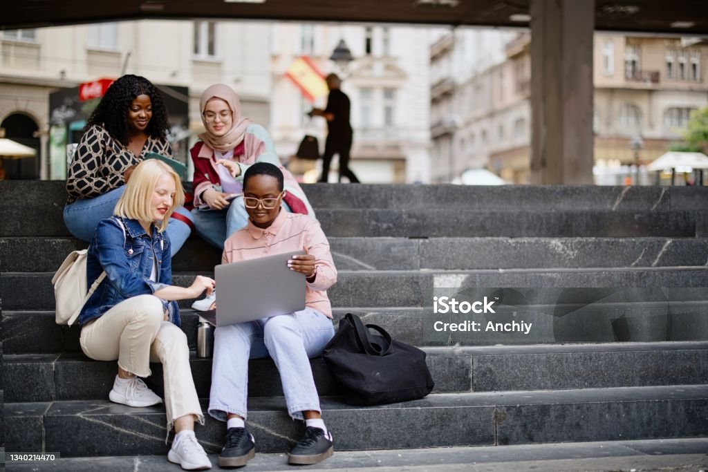 Students sit on the steps near the college and look at the laptop and  digital tablet and talk Four multiethnic girl friends walk around the city after or before the lecture. They wear casual clothes, holding electronic devices such as laptops, digital tablets or smartphones. They discuss lectures, talk and laugh, look at funny memes. University Student Stock Photo