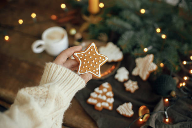 istock Hand in cozy sweater holding gingerbread cookie star on rustic moody background with coffee, fir branches, ornaments on table. Winter hygge. Merry Christmas and Seasons greeting 1340215535