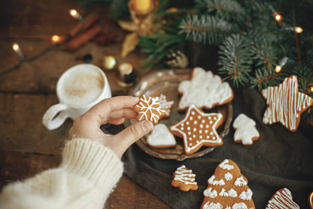 istock Hand in cozy sweater holding snowflake gingerbread cookie on rustic background with fir branches,warm lights, ornaments on table. Winter time. Moody atmospheric image 1340215853