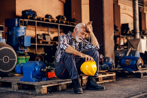 An old, tired bearded factory worker in overalls is sitting on the pallet and taking a break from hard work. He is wiping sweat from his forehead.