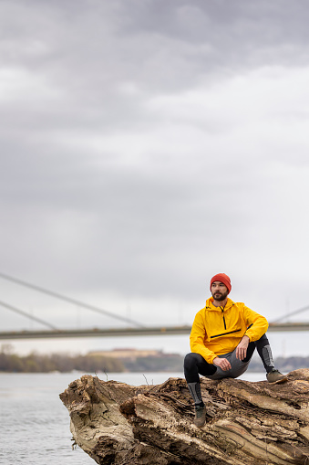 Man sitting on a fallen tree trunk, taking a break while jogging by the river on an overcast autumn day, looking at the distance pensively