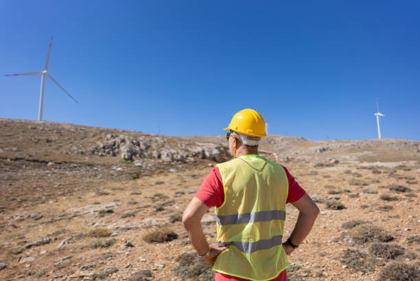 istock Worker or engineer with hard hat and protective clothing looking at a windmill of a wind farm. 1340228875