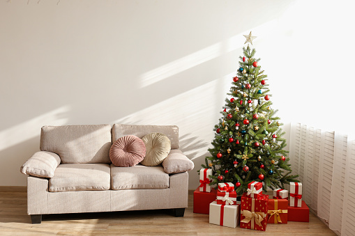 Empty couch and decorated Christmas tree in soft day light from the window of spacious room in lofty apartment. Traditional new year holiday decoration. Close up, copy space.