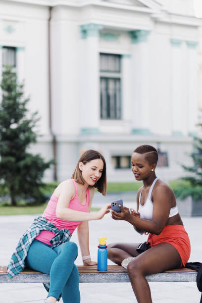 istock Two multiethnic girls relaxing after exercise session, outdoor communicating and using smartphone 1340236350
