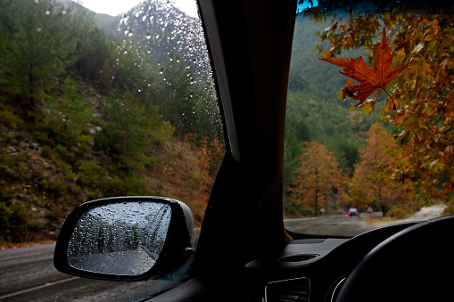 A view out of the car's window with raindrops on the glass. Newly paved switchback road wet after the rain. Autumnal background with yellow and red leaves on trees. Close up, copy space for text.