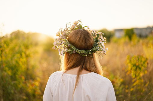 Summer lifestyle portrait of beautiful young woman in a wreath of wild flowers. Standing back in the flower field, hands to the side. Romantic mood. Nature lover