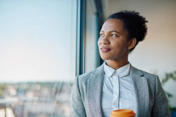 donna d'affari nera che sogna ad occhi aperti mentre beve un caffè e guarda attraverso la finestra dell'ufficio. - looking through window window business women foto e immagini stock