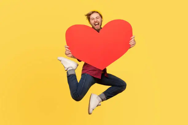 Photo of happy in love man jumping and holding big red heart shape and looking with love and screaming.