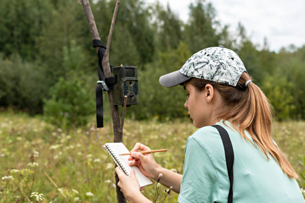 Young woman scientist zoologist writing down data from trap camera to notepad, observing wild animals in reserve taiga forest, monitoring of rare endangered animal species, World Wildlife Day