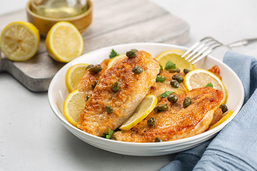 Chicken breast dredged in flour and cooked in sauce cantaining lemon juice, butter and capers. Chicken piccata. White table.