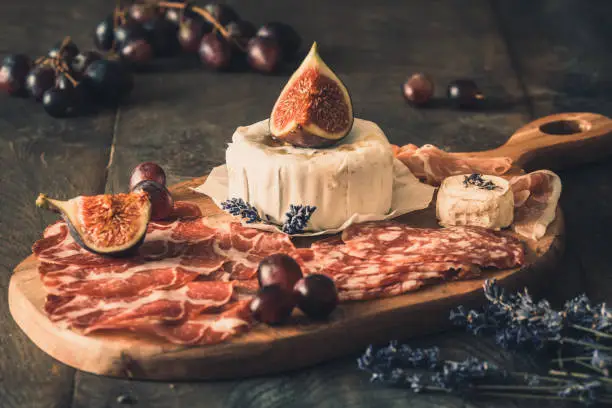 Various smoked sausage specialties and a soft cheese on a wooden board, decorated with figs and grapes