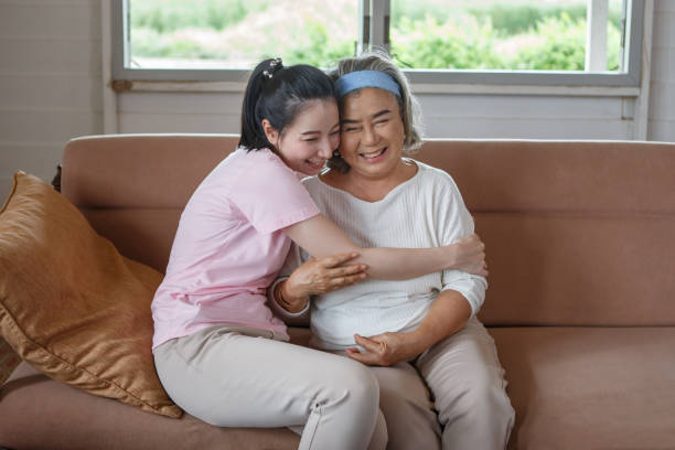 asian adult daughter hug her senior mother on sofa at home . young woman embrace old mom  to take care on couch in living room stock photo
