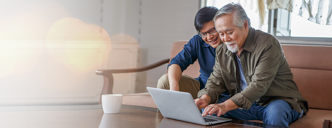 happy asian adult son and senior father sitting on sofa using laptop together at home . young man teaching old dad using internet online with computer on couch in living room . copy space banner