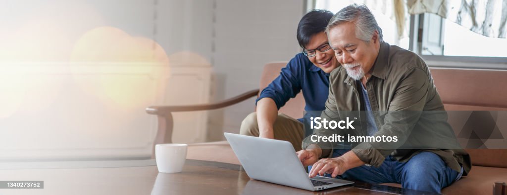 happy asian adult son and senior father sitting on sofa using laptop together at home . young man teaching old dad using internet online with computer on couch in living room . copy space banner Senior Adult Stock Photo