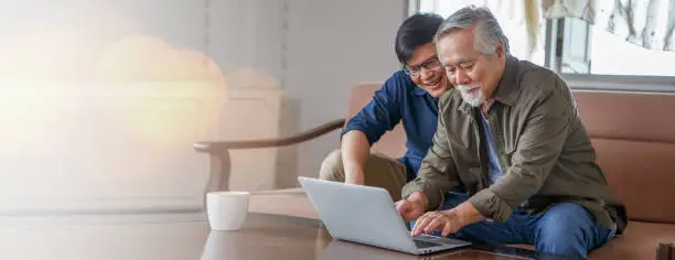 Photo of happy asian adult son and senior father sitting on sofa using laptop together at home . young man teaching old dad using internet online with computer on couch in living room . copy space banner