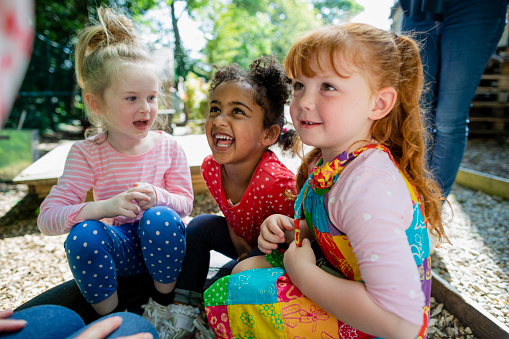 A low angle close up of a little girl and her friends sitting in the school playground and playing and having fun together and enjoying being children. Their teacher is there also to supervise and make sure they behave.