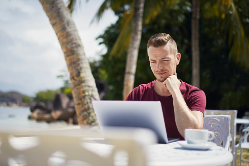Young freelancer working on laptop from tropical destination. Man sitting under palm trees on beach.