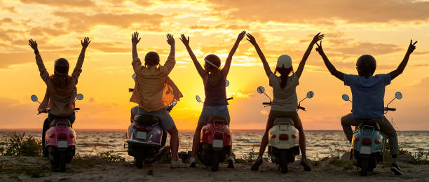 istock Back view of cheerful young friends, motorcycle riders raised their hands while standing with their scooters and watching sunset together 1340294010