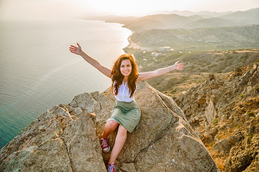 Panoramic photo of a happy woman with open hands who conquered the top of a cliff with a beautiful view of the sea and the cityscape. The concept of travel and freedom.