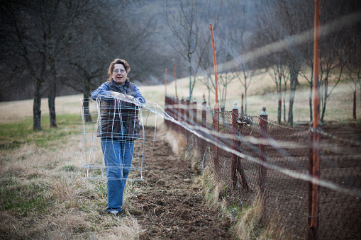 Woman farmer building and repairing protective fence of vegetable garden in rural countryside