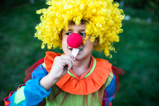 A cute boy in a clown costume is trying to bile off a piece of chewy Halloween candy