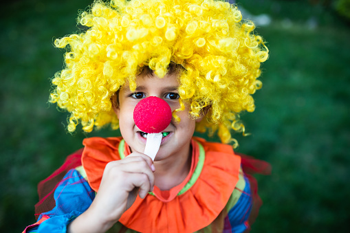 A cute boy in a clown costume is trying to bile off a piece of chewy Halloween candy