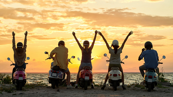 Silhouette of cheerful relaxed people, motorcycle riders wearing helmets, raised their hands while standing with their scooters and watching sunset together. Friendship, nature concept. Rear view