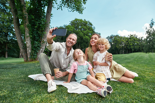 Lovely family spending time together outdoors, using smartphone while taking a selfie, having picnic in nature on a summer day. Leisure, summer, technology concept