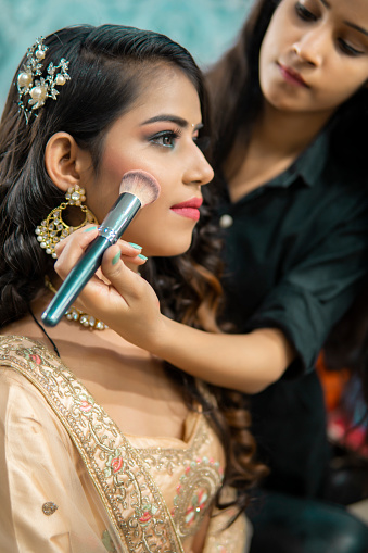 Female makeup artist doing makeup of a beautiful Indian woman for a wedding reception at the beauty parlor.