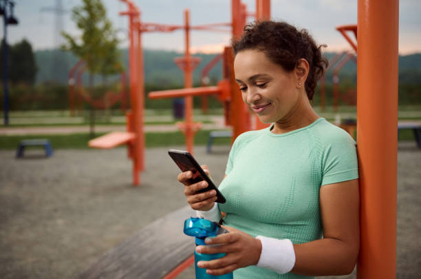 attractive athlete mixed race beautiful young woman holding a bottle with water and checking mobile application on her smartphone, resting after sport activities outdoors - playing field effort outdoors human age imagens e fotografias de stock