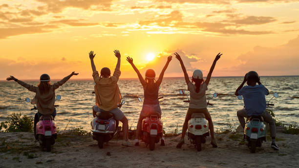 Back view of young friends, motorcycle riders raised their hands while standing with scooters on the beach and enjoying sunset together