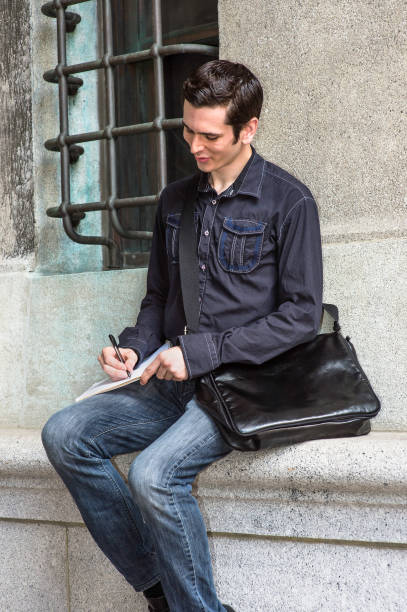 istock Young man sitting by wall in New York City, writing on notebook 1340308842