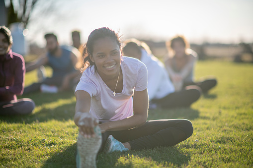 A group of young ethnically diverse friends workout together in a park. They are smiling as they are seated with one leg laying out and their arms stretched to the tip of their toes.