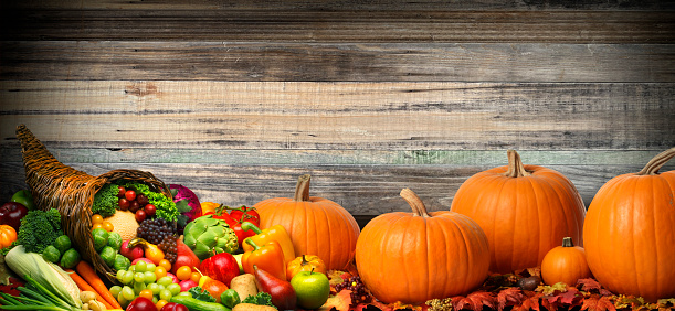 A Thanksgiving cornucopia of fruits and vegetables and a group of harvest pumpkins rest on a table in front of a background of wooden planks.