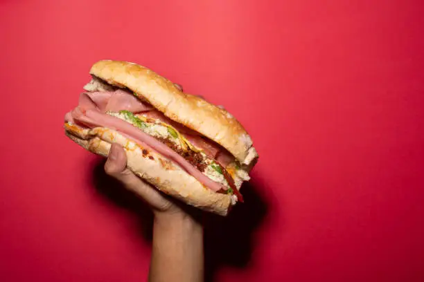 a hand holds a barda cake, mexican cake, mexican food, mexican sandwich with chiruzo, ham, avocado, white cheese, ceboola, tomato on a french bread, bolillo bread