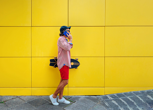 a man is carrying a skateboard while walking and talking on the phone. the background is yellow.