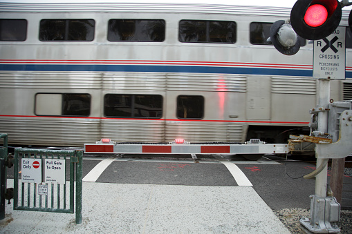 silver double deck train with red and blue strip. Crossing arm down. Stop light is red. Train is slightly blurred as speeding by