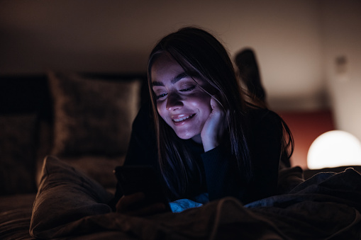 Bright and happy smiling Young Woman lying in her Bed in Bedroom at Night, reading good and funny news on her mobile phone. Laughing and smiling bright over the funny messages. Ambient Night Bedroom Light. Millenial Generation Modern Technology Lifestyle.