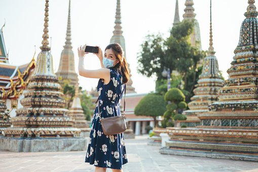 Young thai/chinese social distancing safety travel new normal lifestyle asian female woman wearing protective face mask walking and take photo with smartphone tour in ancient asia temple in thailand sunset moment