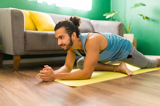Cheerful latin man practicing a pigeon pose on an exercise mat in his living room. Attractive man exercising with yoga