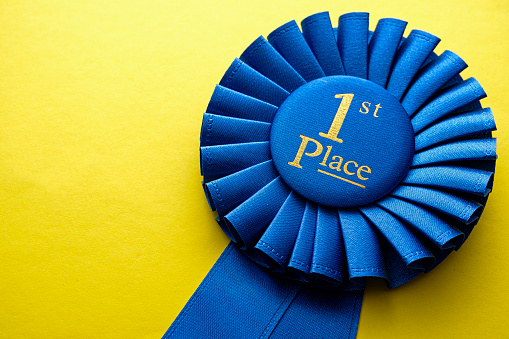 First place winners rosette with blue ribbon and gold text on a yellow background with copyspace