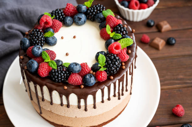 istock Three chocolate mousse cake with chocolate glaze, blueberries, raspberries and blackberries on top on a white plate on a dark wooden background. Cake with three layers of different taste. Copy space. 1340425963