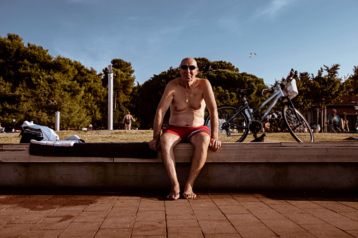 Cheerful local senior man, sitting on the bench near the beach, sunbathing and observing the environment